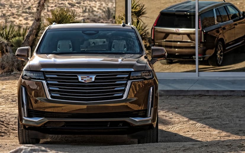Bronze Cadillac Escalade luxury SUV stands in a desert beside a glass wall reflecting its rear profile.