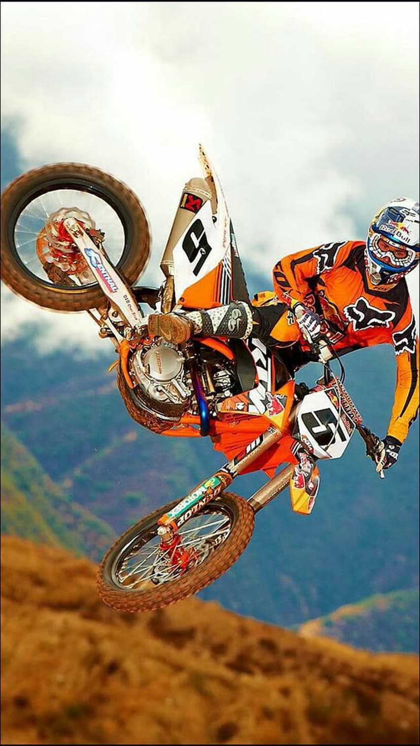 Motocross athlete in gear performs a high dirt bike jump against a cloudy sky and blurred mountains.