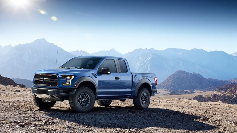 Blue Ford F-150 Raptor truck on rocky desert ground with sunlit mountains and a clear blue sky background.