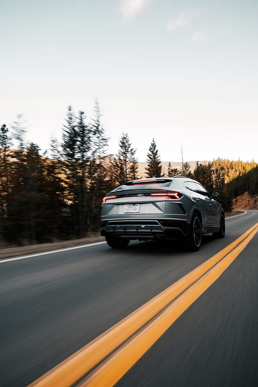 Silver SUV with glowing red taillights cruises through a pine forest road during a vibrant mountain sunset.