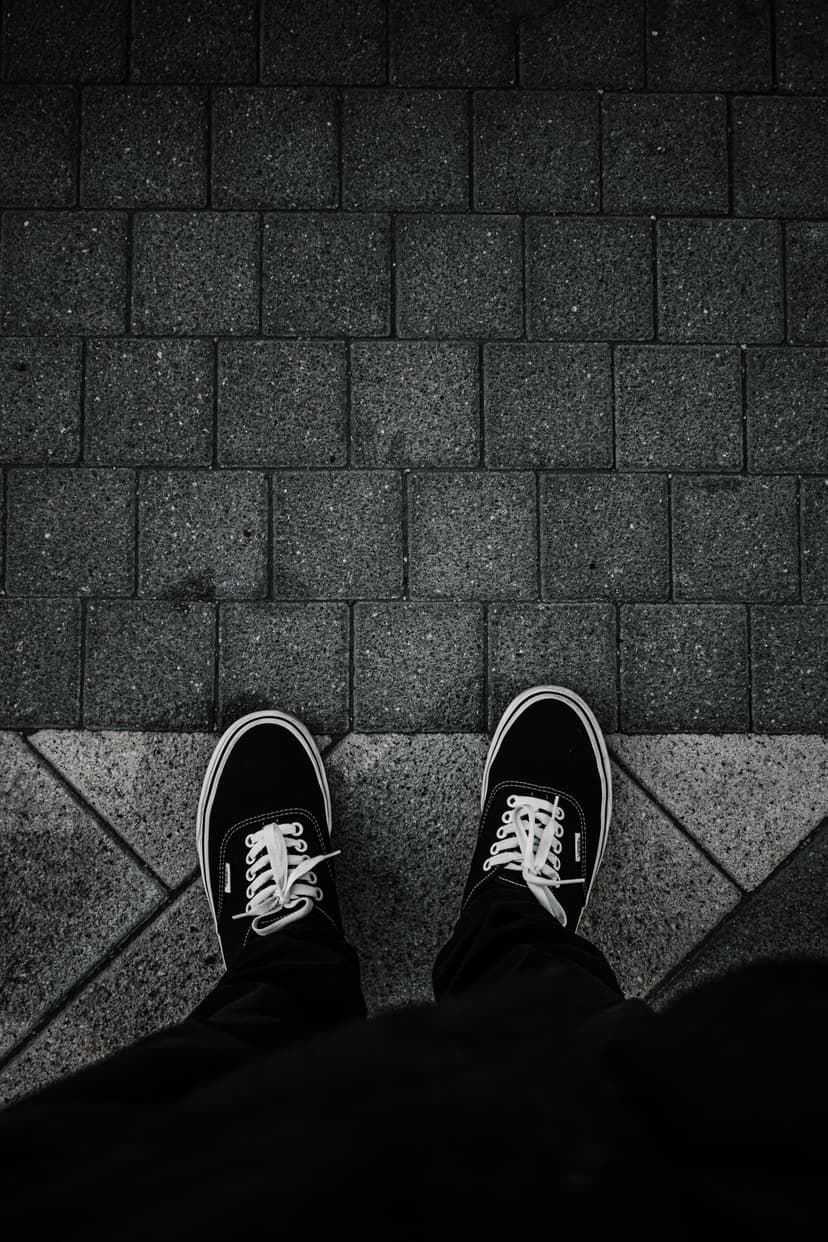 Black canvas sneakers with white laces rest on a geometric gray stone pavement in a vertical top-down view.