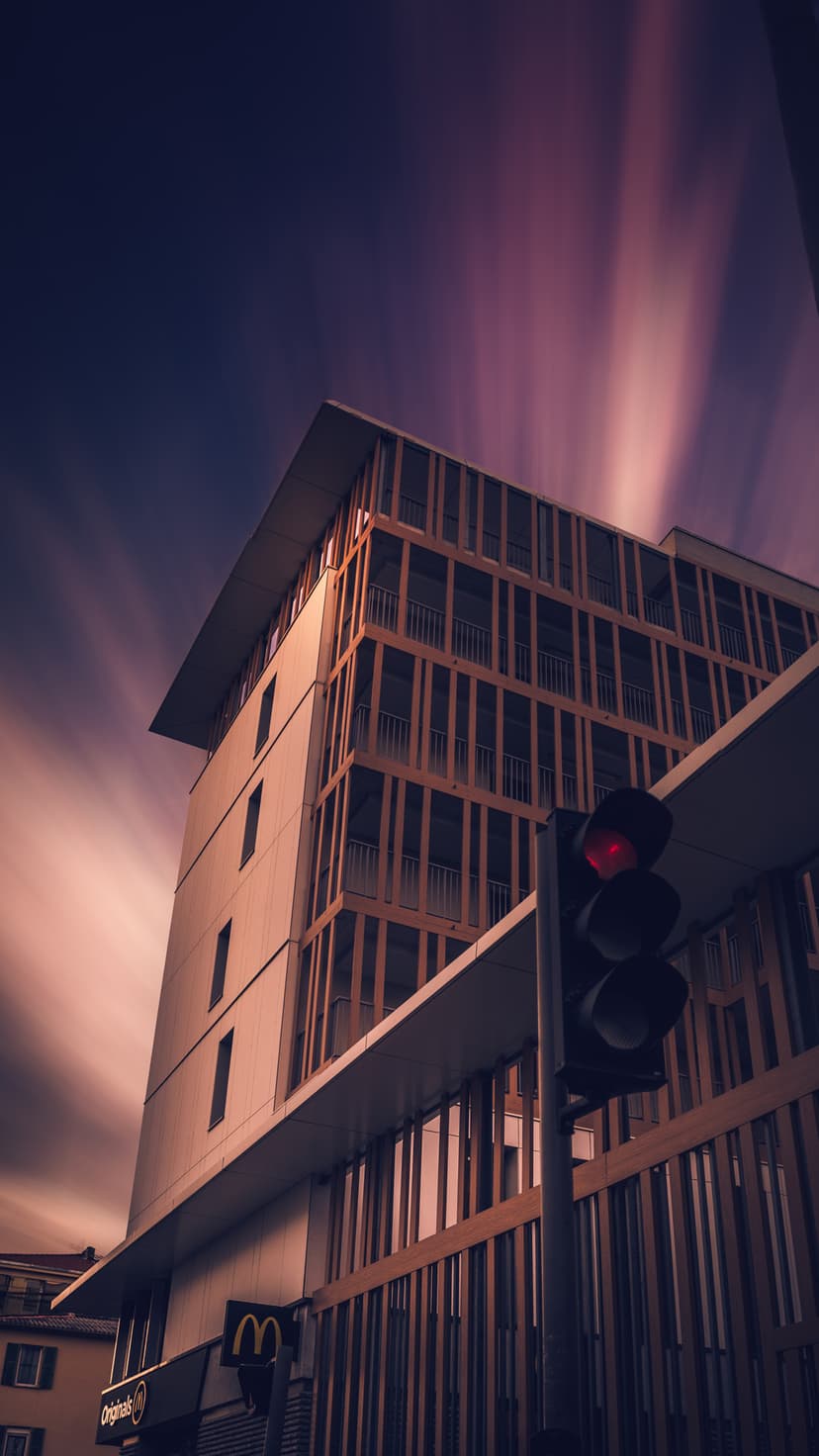 A tall modern building with a geometric facade stands beneath long exposure cloud streaks and a red street light.