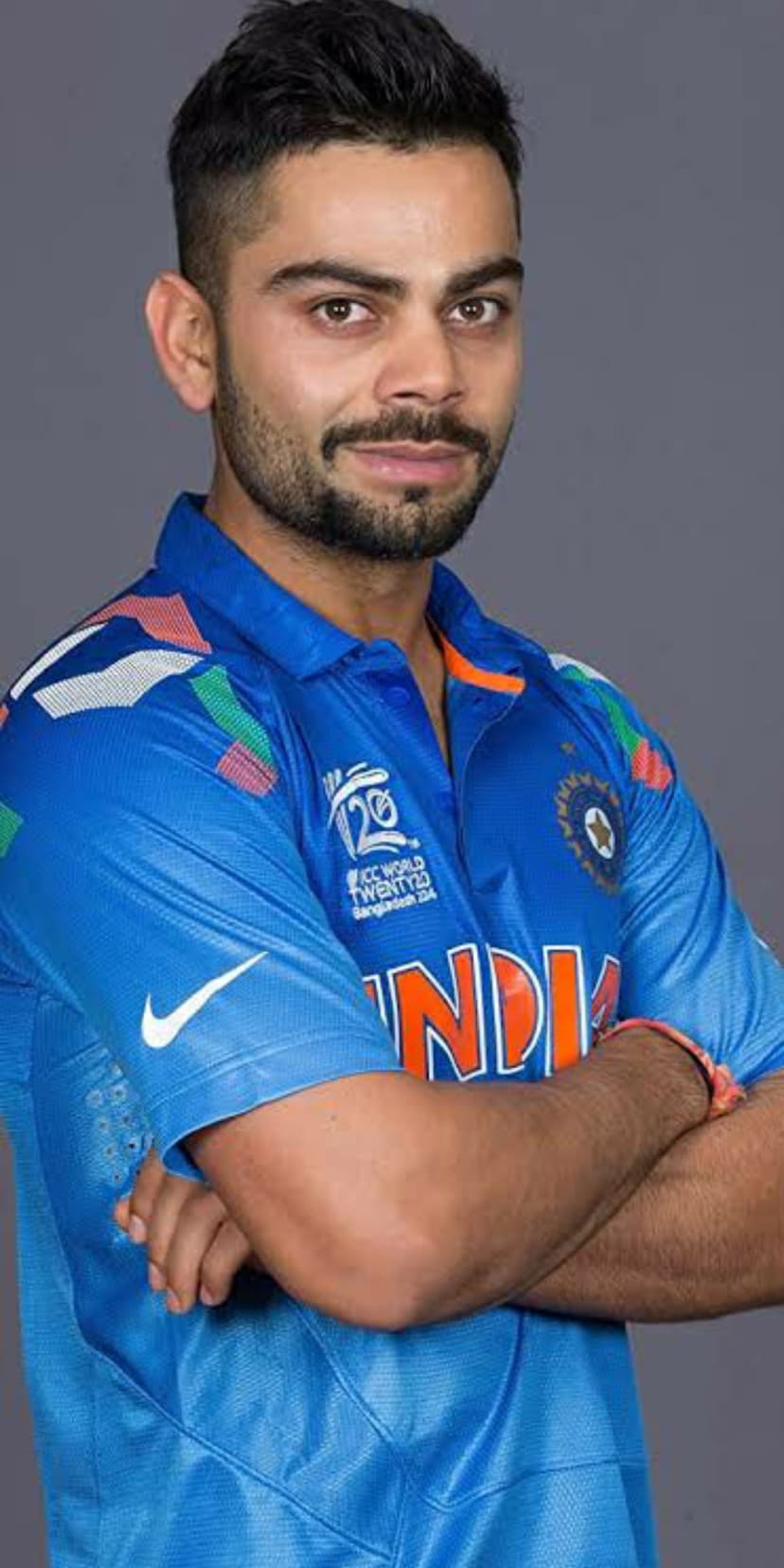 Virat Kohli wearing a blue India cricket jersey with arms crossed and a confident expression in a studio portrait.