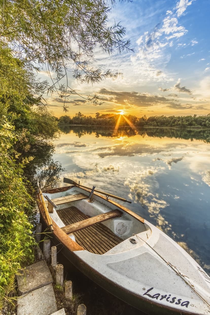 Wooden rowboat named Larissa docks near a grassy shoreline under a vibrant orange and gold sunset sky.