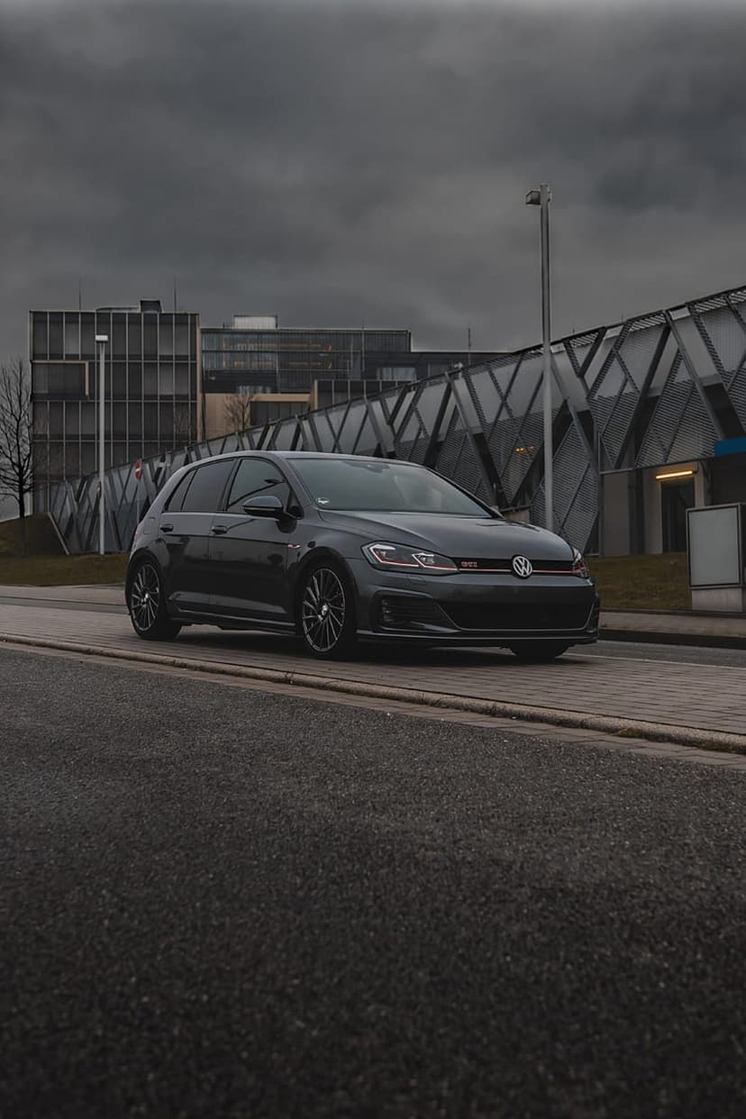 Dark gray Volkswagen Golf GTI parked on a city street near a modern building under a dramatic overcast sky.