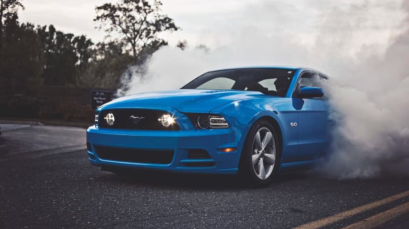 Blue Ford Mustang GT performing a burnout with thick white smoke on a dark asphalt road under a cloudy sky.