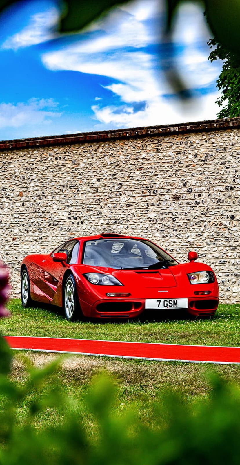 Red McLaren F1 supercar parked on green grass beside a textured stone wall under a blue sky with white clouds.