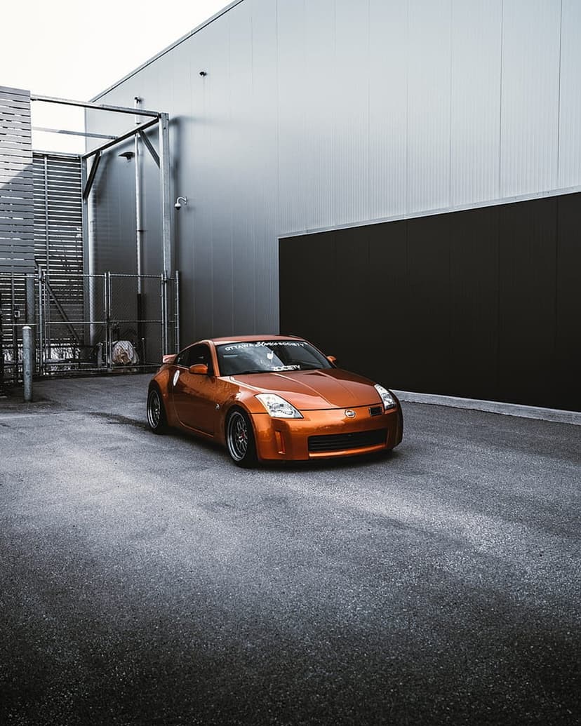 Orange Nissan 350Z sports car parked on asphalt in front of a corrugated metal industrial building facade.