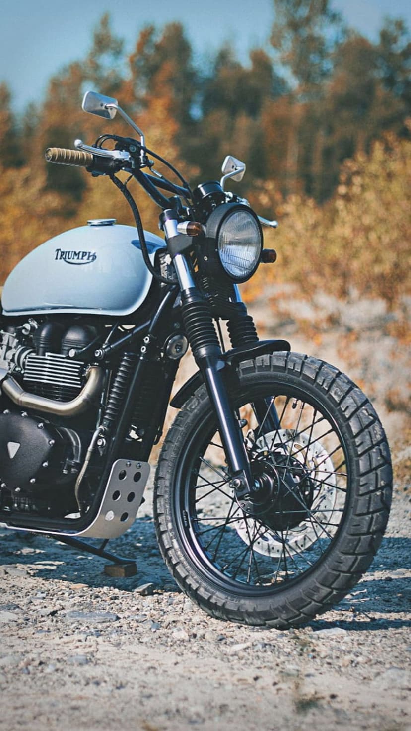 Blue Triumph Scrambler motorcycle with knobby tires parked on a gravel road surrounded by autumn trees.