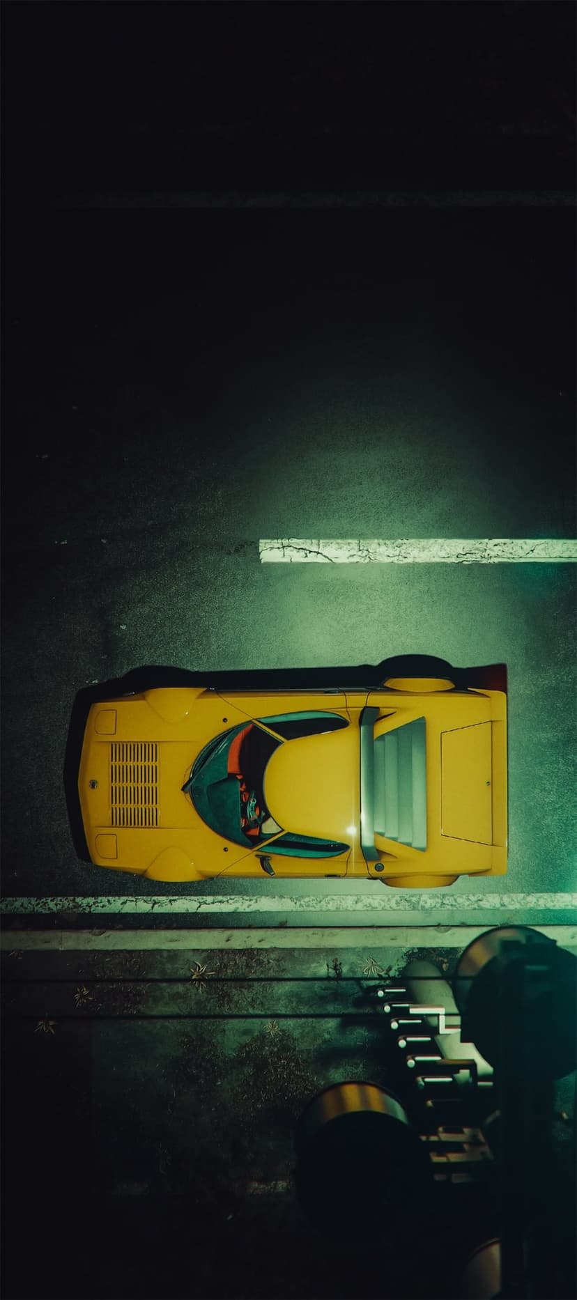Yellow Lancia Stratos style sports car parked on a dark, textured asphalt road seen from an aerial perspective.