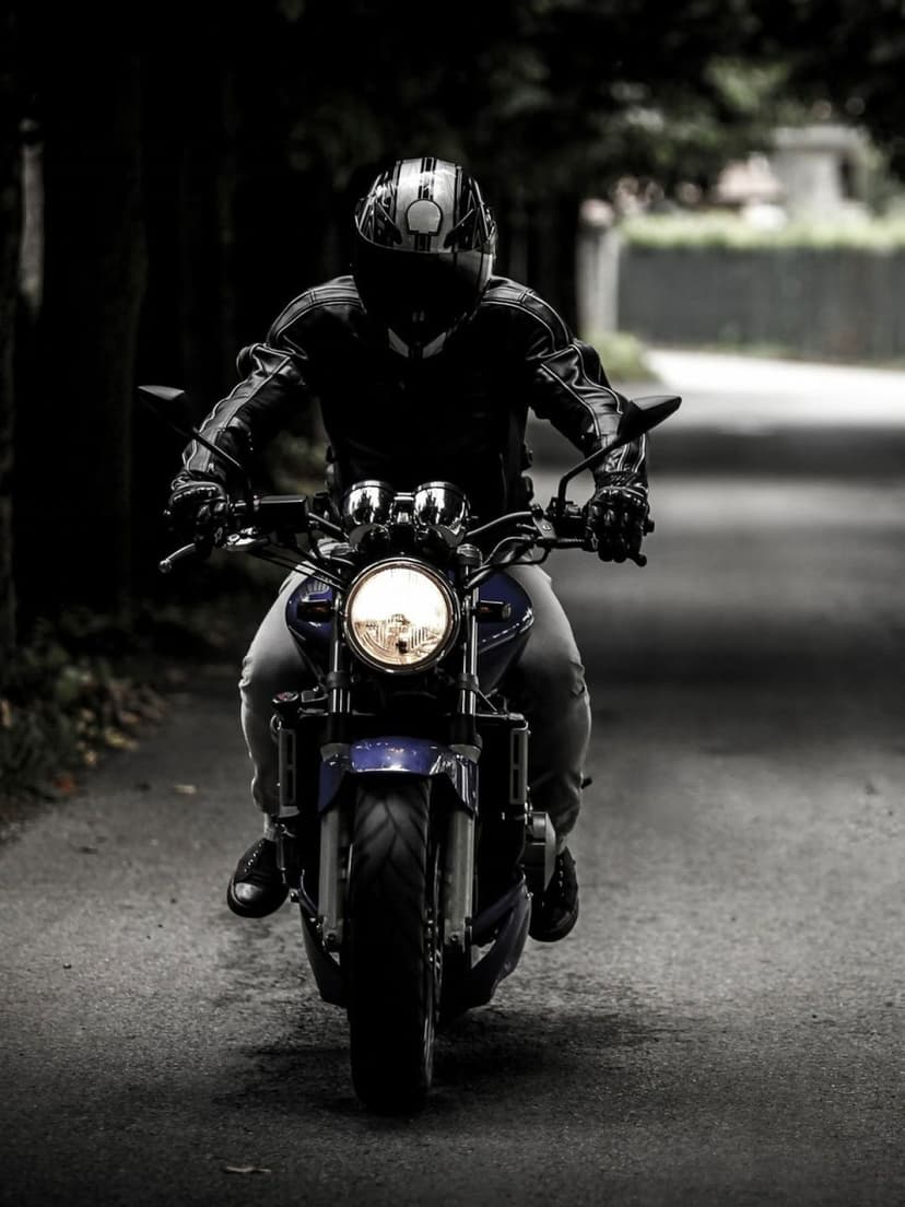 Motorcyclist in a leather jacket riding a blue bike on a dark tree-lined road with a glowing headlight at dusk.
