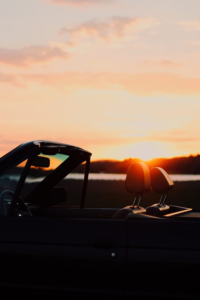 Black convertible car interior silhouette overlooking a pink and orange sunset sky and a distant lake.