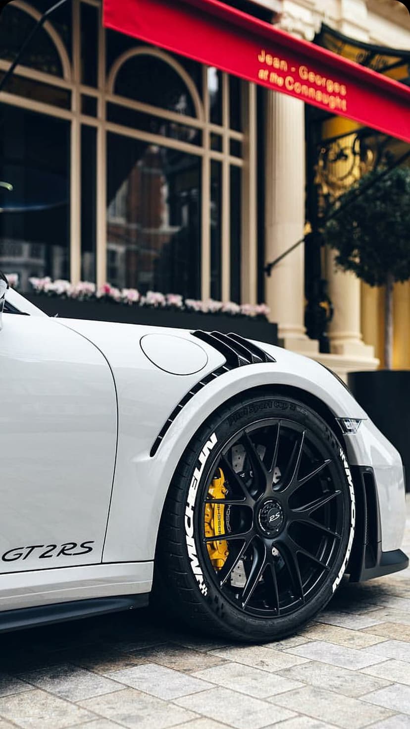 White Porsche GT2 RS features black multi-spoke wheels and yellow brake calipers against an urban building.