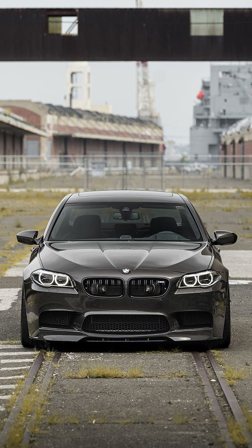 Dark gray BMW M5 sedan front view parked on weathered railroad tracks with blurred industrial cranes in the distance
