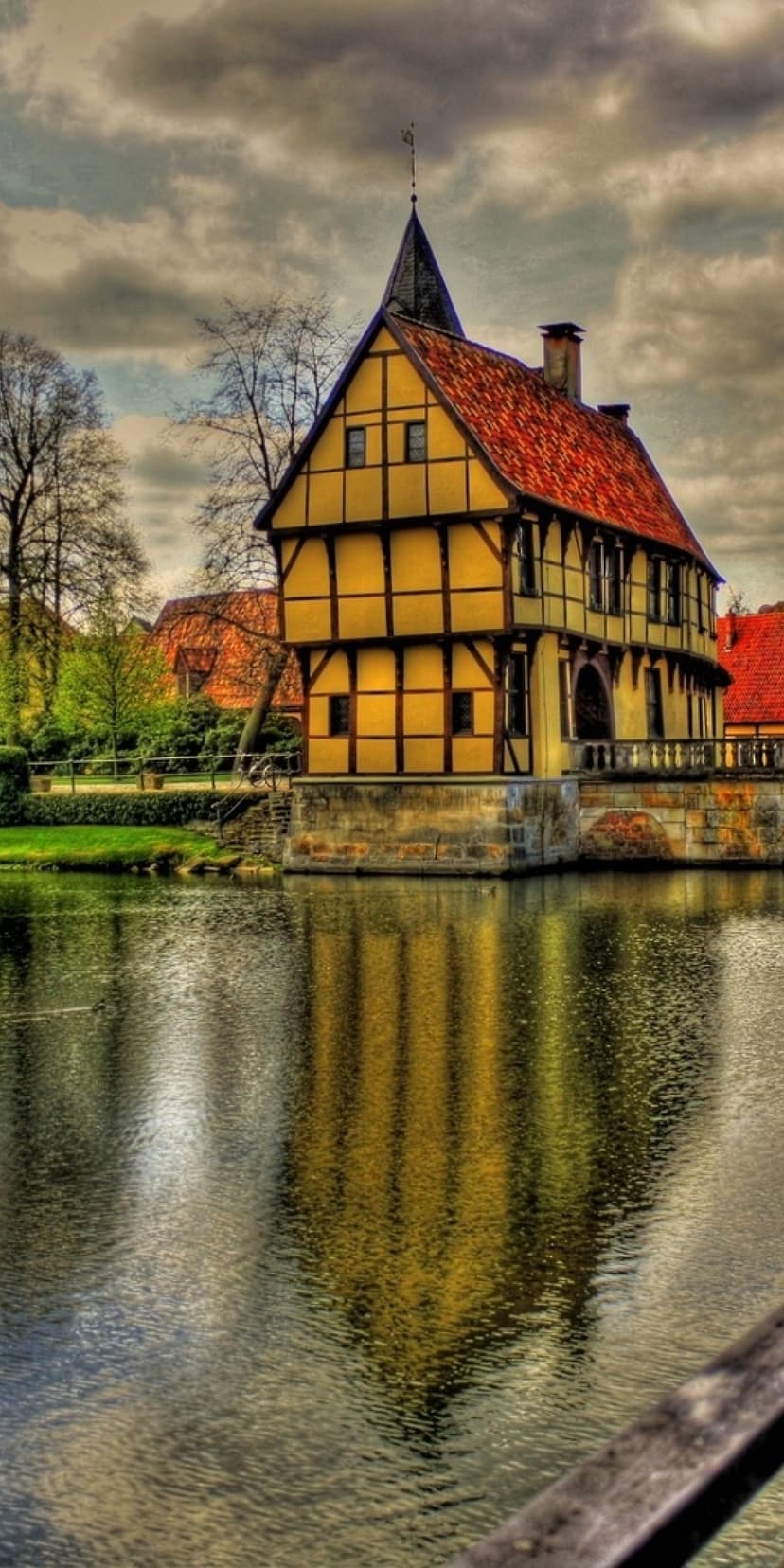 Yellow half-timbered house reflects on a calm lake surface with green trees and a cloudy sky in the background.