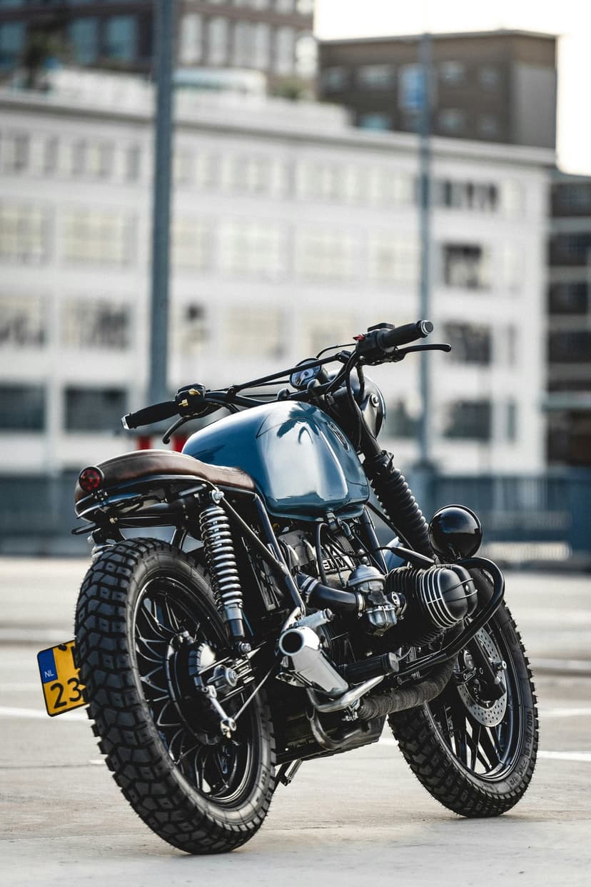 Blue cafe racer motorcycle featuring knobby tires and a brown seat parked outdoors near a modern building.
