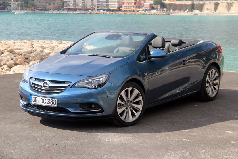 Blue Opel Cascada convertible parked on a coastal road beside turquoise sea water and town buildings.