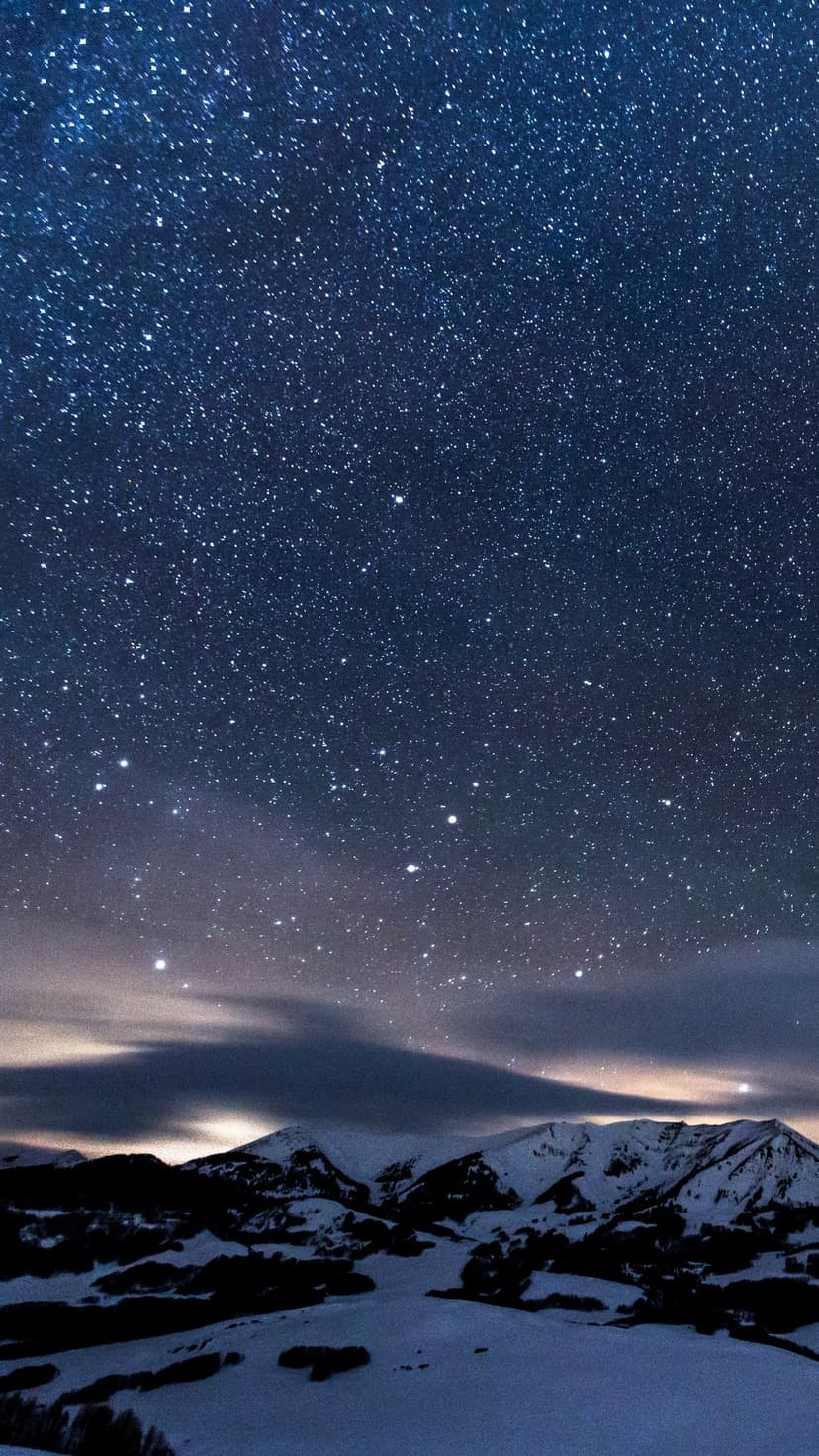 Star-filled night sky over sharp snow-covered mountain peaks under a vast cosmic galaxy in a winter wilderness.