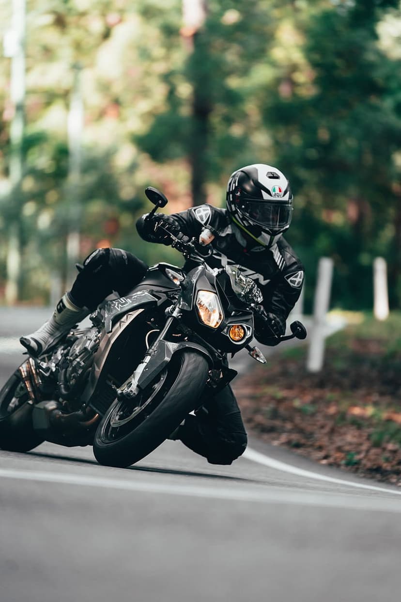 Motorcyclist in full gear leans a sportbike into a sharp turn on a paved road surrounded by dense green trees.