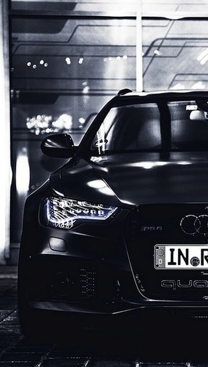 Audi RS6 front grille and glowing LED headlights emerge from shadows against a textured modern wall.