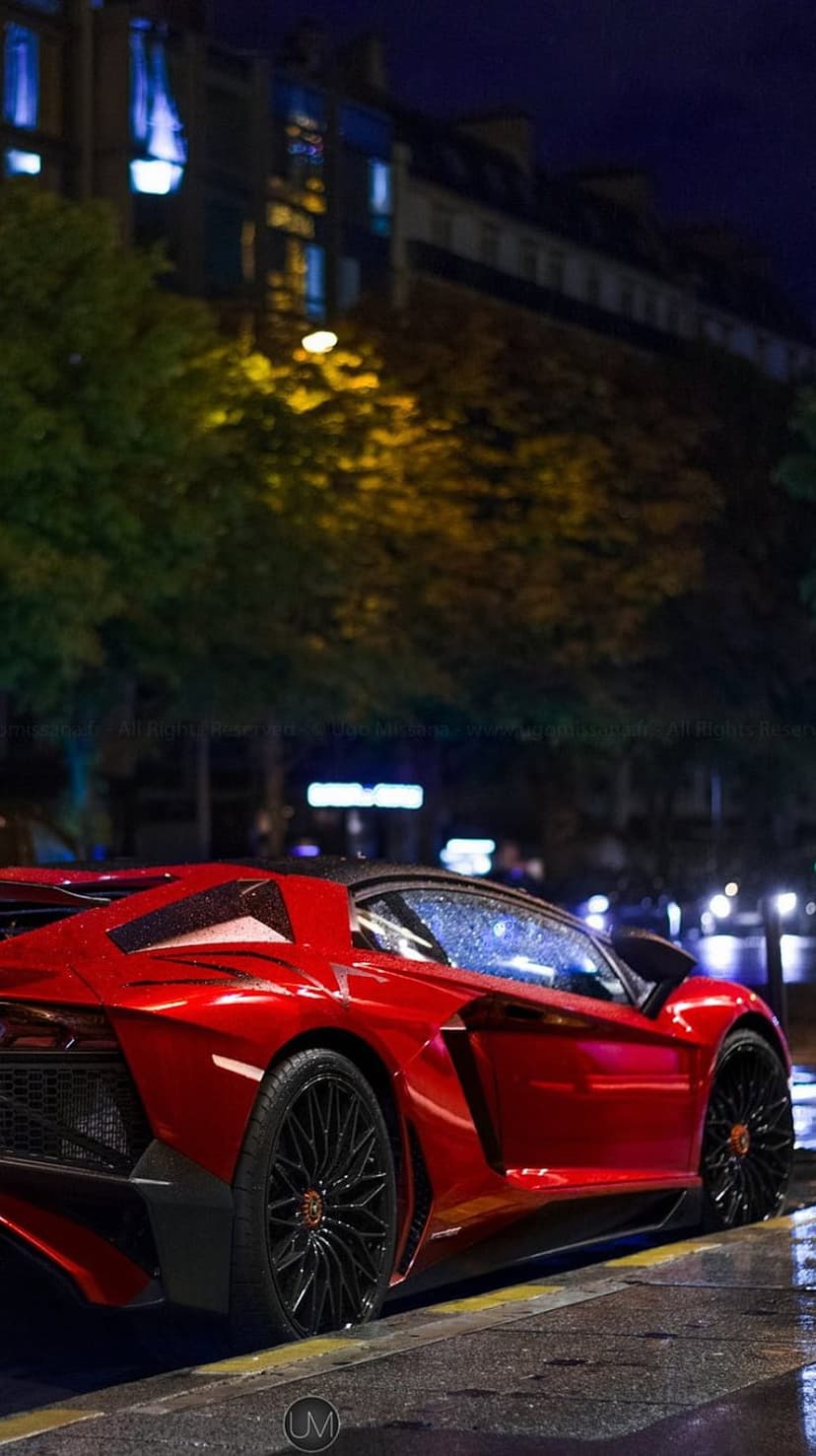 Red Lamborghini Aventador with raindrops on its hood parked on a wet urban street under nighttime city lights.
