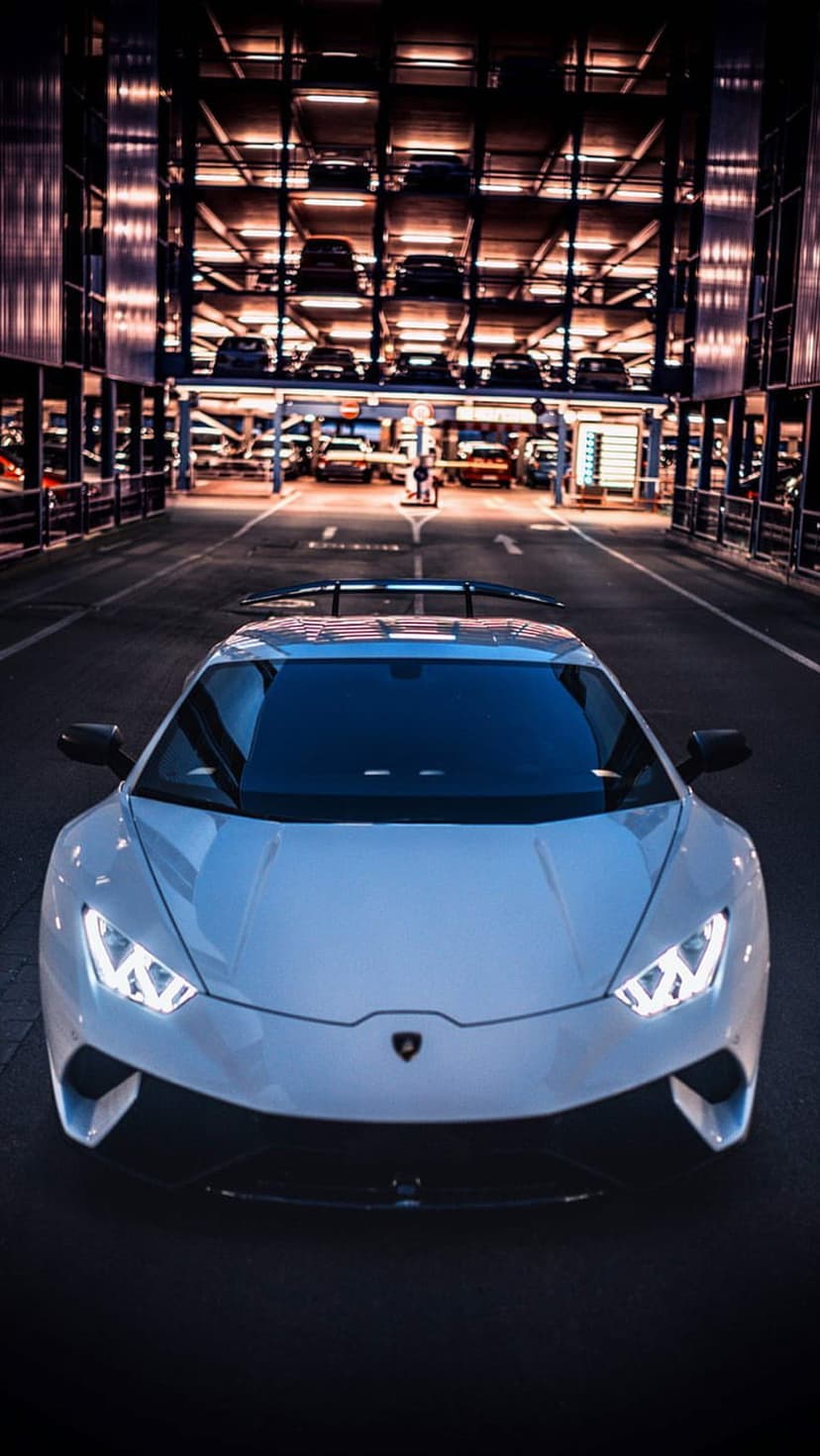 White Lamborghini with glowing headlights parked in a dark multi-level garage under warm ambient lights.