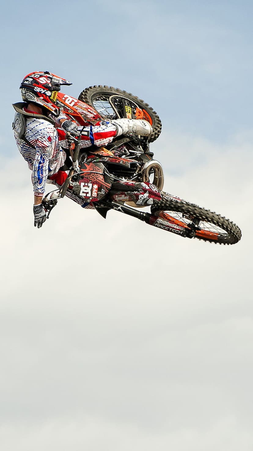 Motocross rider in a red and blue suit jumps a black and red dirt bike high against a pale cloudy sky background.