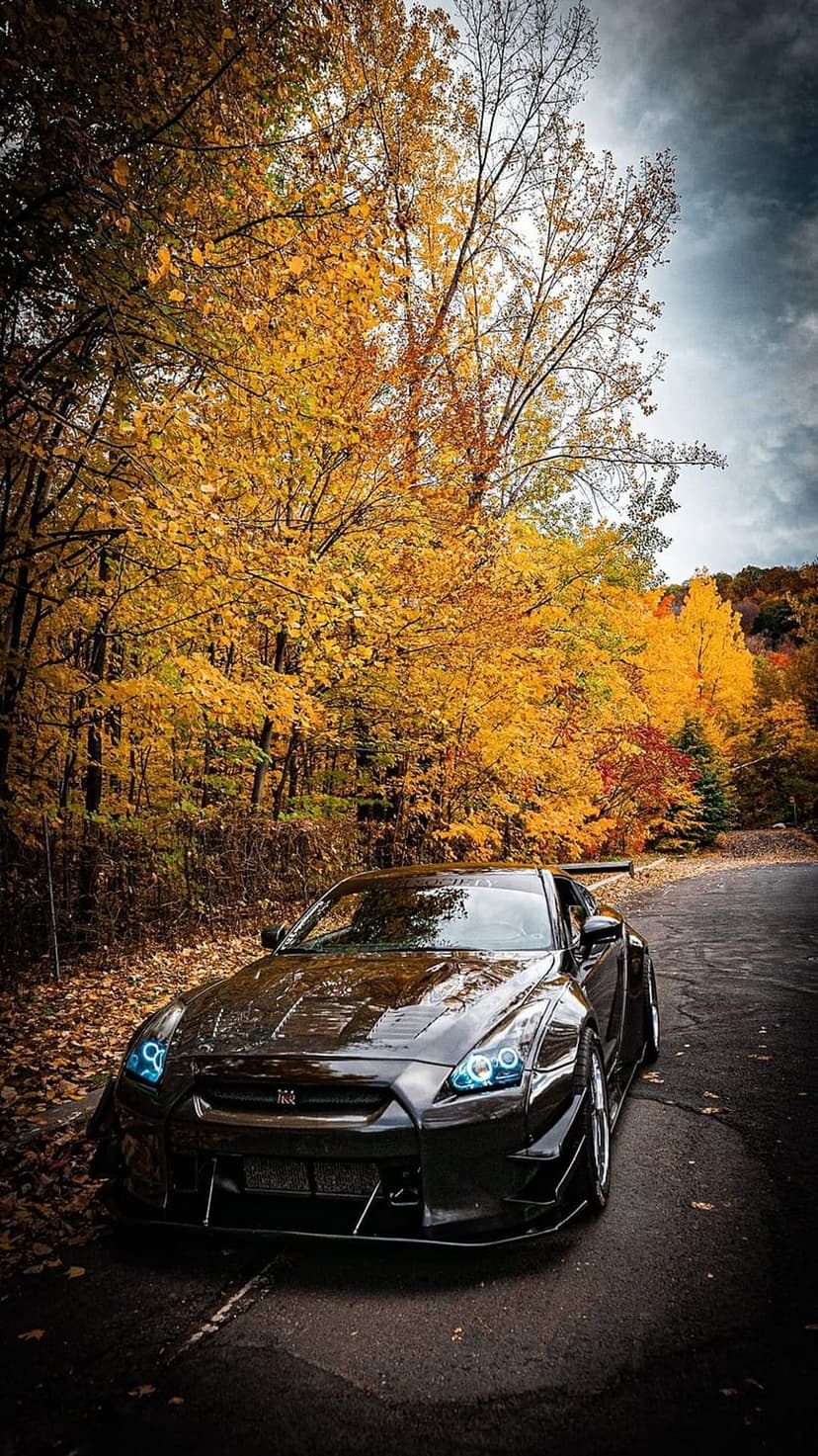 Black Nissan GT-R parked on a winding forest road surrounded by yellow autumn leaves and moody clouds.