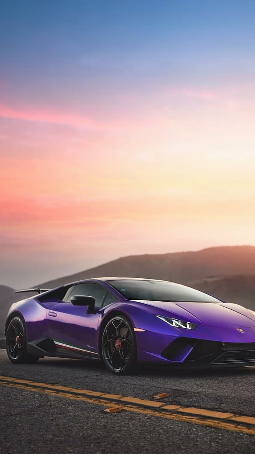 Purple Lamborghini Huracan drives on a winding road against a vibrant orange and pink sunset sky with distant hills.