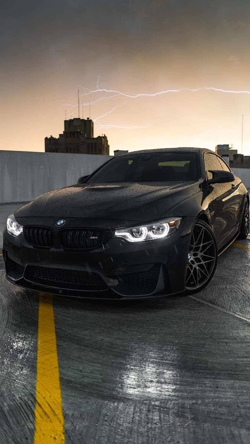 Black BMW M4 with glowing headlights parked on a wet rooftop during a lightning storm under an orange sky.