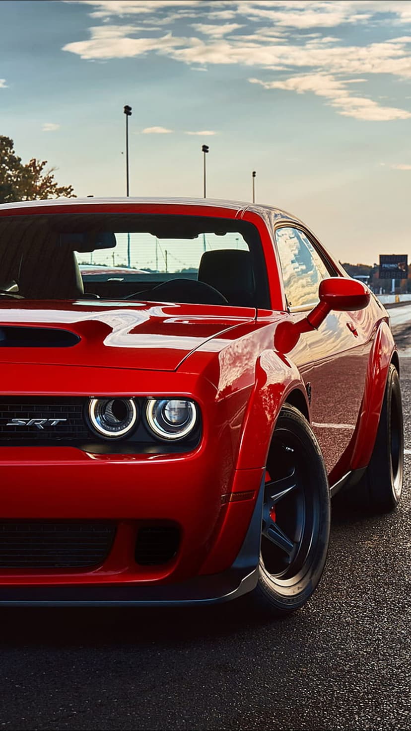 Red Dodge Challenger SRT Demon with glowing halo headlights parked on a dark asphalt track during a warm sunset.