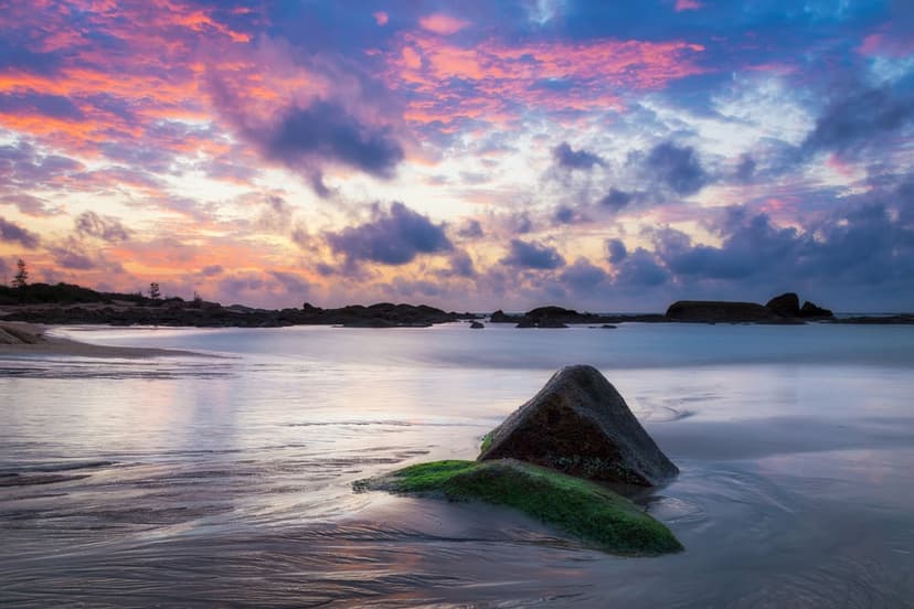 Mossy rocks sit on a wet sandy beach under a vibrant purple and pink sunset sky during a long exposure.