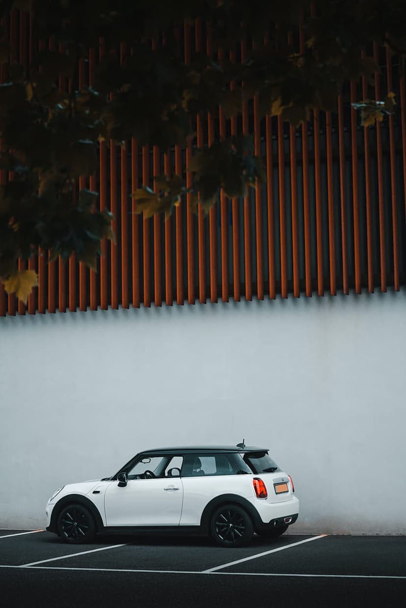 White Mini Cooper parked against a white wall and orange slatted building with autumn leaves at the top frame.