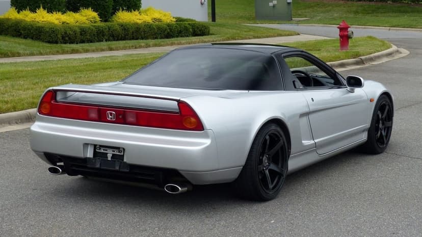 Silver Honda NSX sports car with black wheels and a continuous red tail light bar parked on a grey asphalt road.