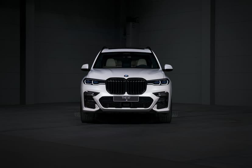 Front view of a white BMW X7 with glowing blue headlights centered in a dark, shadowy minimalist studio setting.