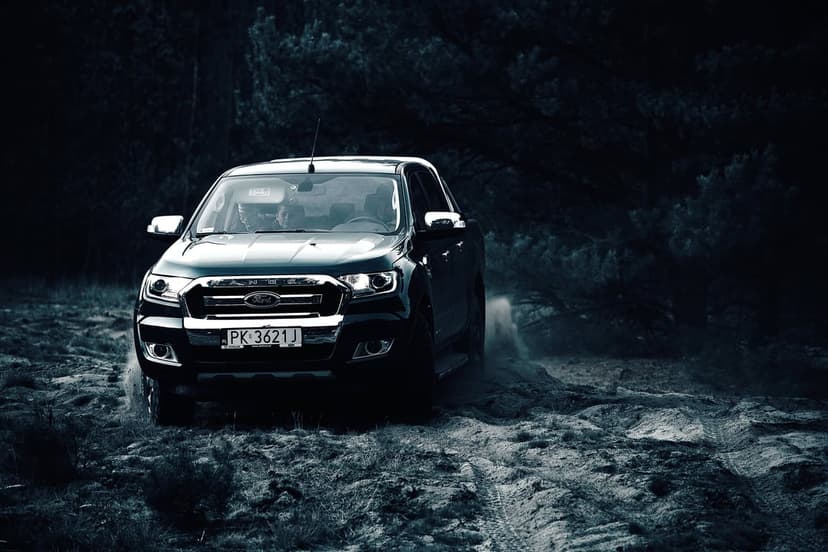 Ford Ranger truck with glowing headlights driving through a dusty dark forest on a rugged dirt path at night.