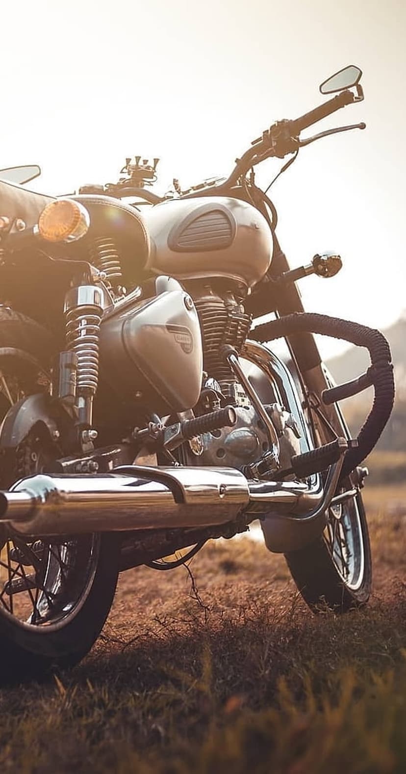 Classic motorcycle engine and chrome exhaust pipe resting in dry grass during a hazy golden hour sunset.
