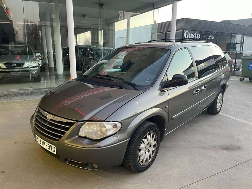 A grey Chrysler Voyager minivan parked on concrete in front of a modern glass building under soft light.