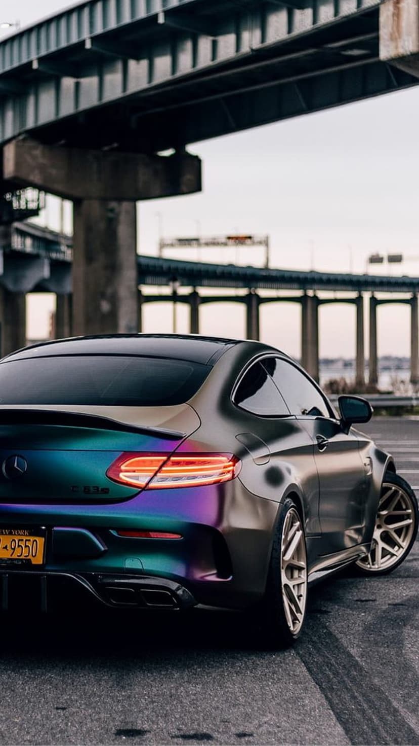 Mercedes-AMG C63S with iridescent purple and teal wrap parked under a concrete bridge with red brake lights on.