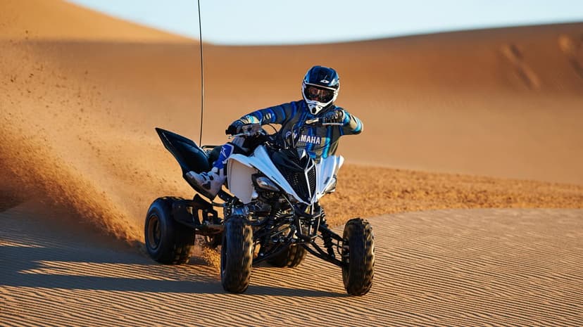 A rider on a white Yamaha ATV power slides across textured sand dunes under a clear blue desert sky.