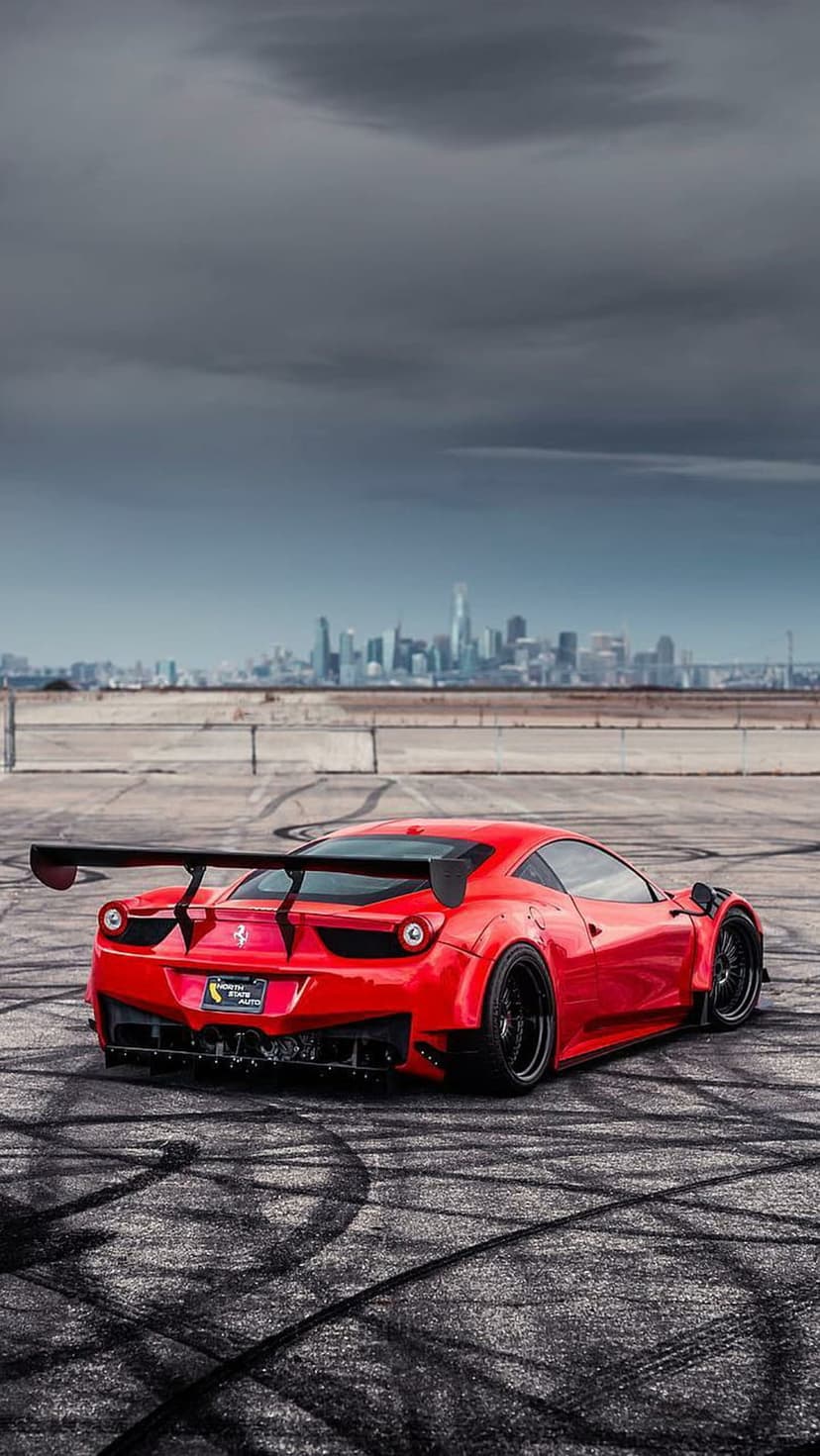 Red Ferrari 458 Italia with a massive rear wing and widebody kit sits on a race track with a city background.