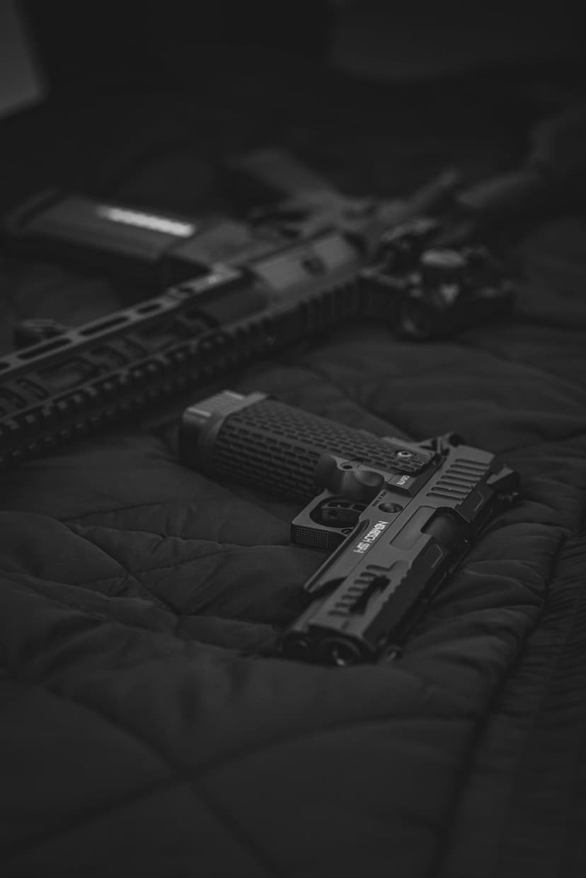 Tactical handgun with textured grips and a blurred rifle rest on a dark, moody grayscale metallic surface.