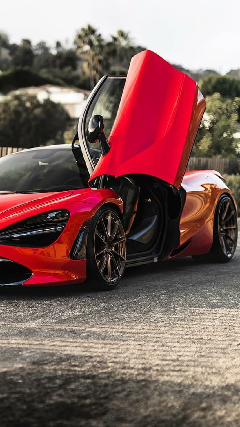 Red McLaren supercar with butterfly doors open on an asphalt road with a blurred green forest background.