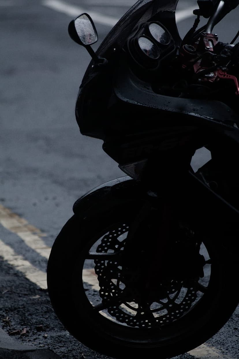 Motorcycle front wheel and brake disc silhouetted against a wet grey road with yellow lines and rain reflections.