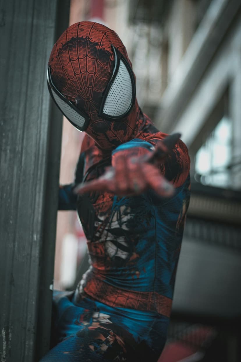 Spider-Man cosplayer in a red suit leans against a wall while extending an outstretched hand in a city.