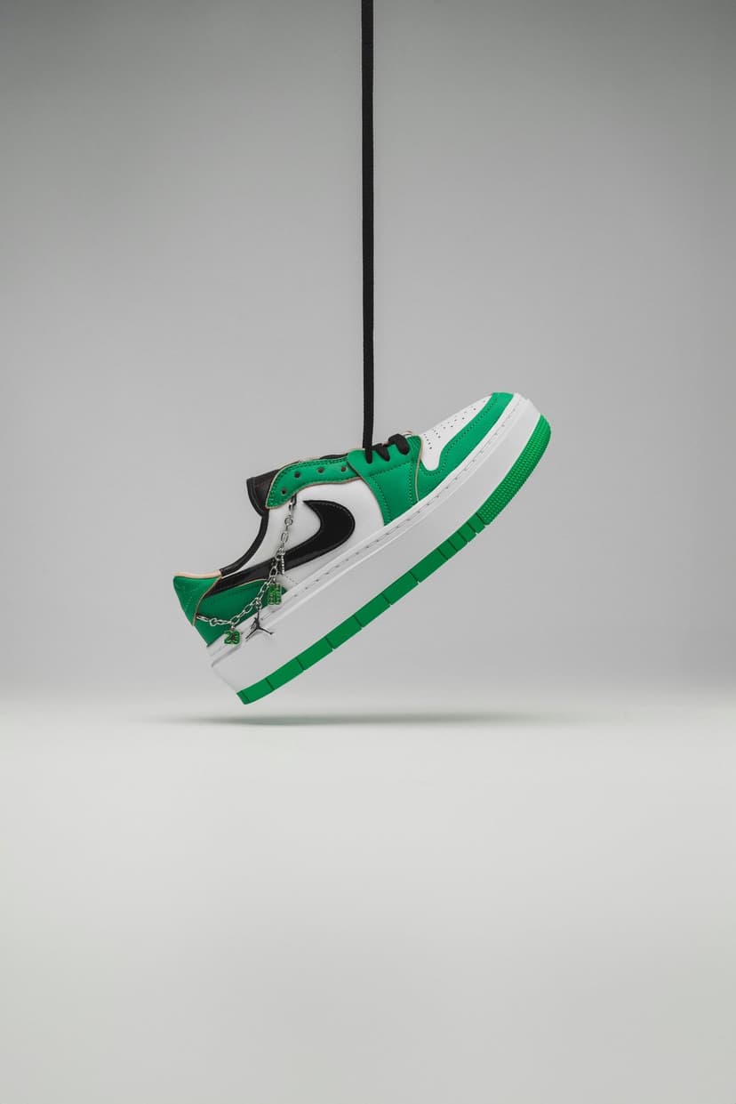 Green Nike Air Jordan 1 platform shoe hangs by a black cord with silver charms against a gray studio backdrop.