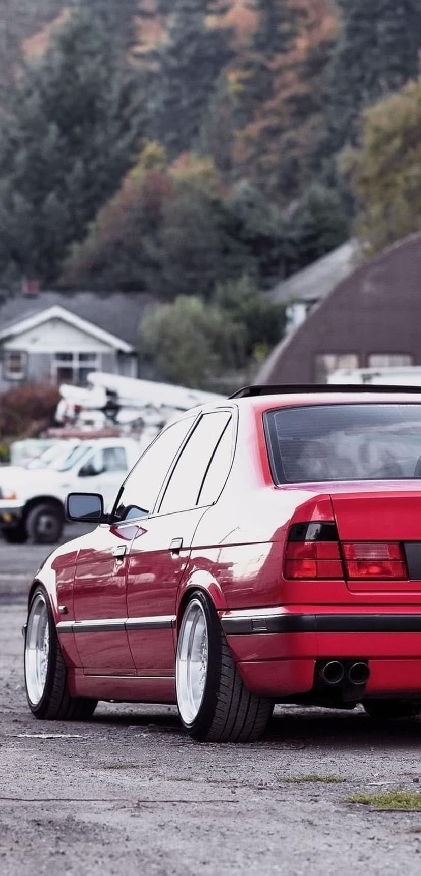Red BMW sedan with polished chrome wheels parked on gravel against a suburban backdrop of green trees and houses.