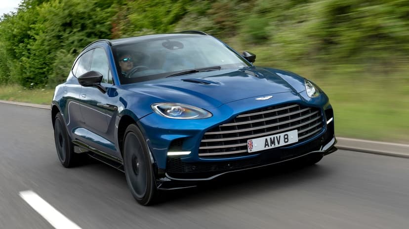 Blue Aston Martin SUV driving fast on a paved road with blurred green trees in the background.