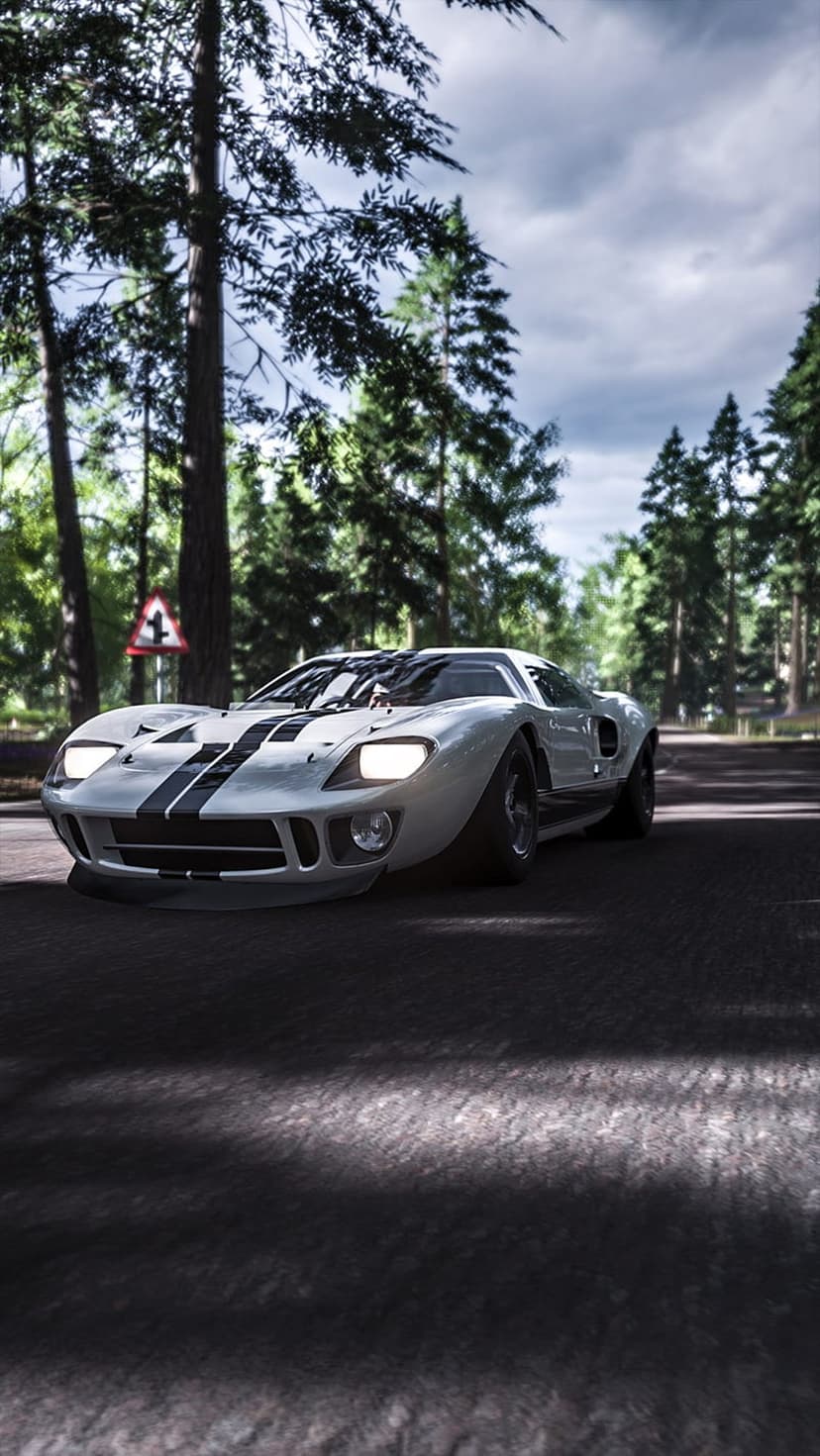 White Ford GT40 with black stripes racing on a paved forest road under a moody, overcast grey sky.