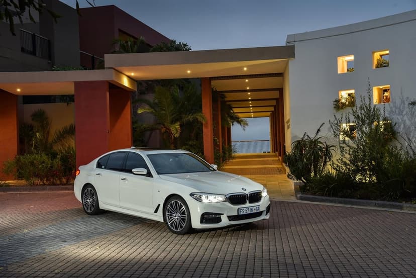 White BMW sedan parked on a brick driveway at dusk in front of a modern house with warm glowing lights.